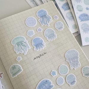 May include: A sheet of stickers featuring watercolor-style illustrations of jellyfish in shades of blue and green. The stickers are arranged on a gridded notebook page with the text "jellyfish" written below them.