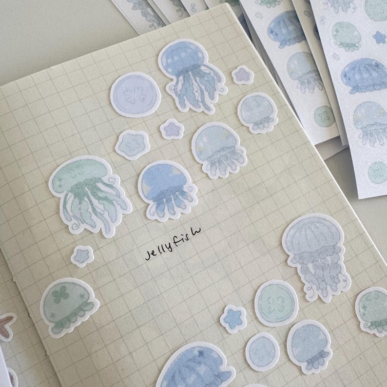 May include: A sheet of stickers featuring watercolor-style illustrations of jellyfish in shades of blue and green. The stickers are arranged on a gridded notebook page with the text "jellyfish" written below them.