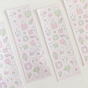 May include: Four sheets of pink sticker paper with a repeating pattern of white rabbits holding four-leaf clovers. The stickers are surrounded by small pink flowers and green clovers. The text on the sticker sheet reads "©2024 dear mini".