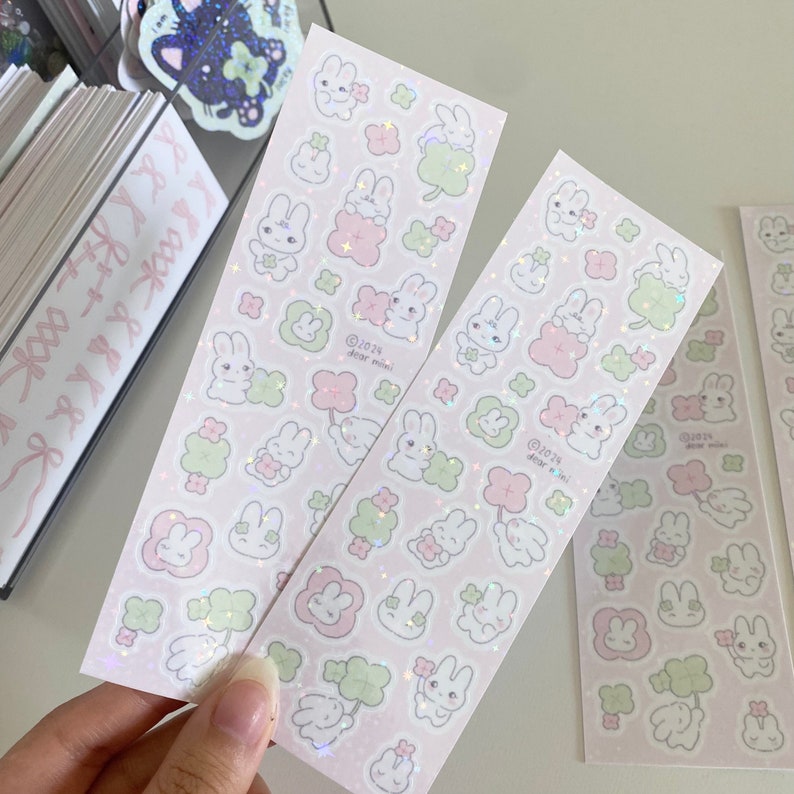 May include: Two sheets of pink holographic stickers featuring cute cartoon bunnies holding green four-leaf clovers. The stickers are decorated with glitter and the text "@2023 dear mimi!" is printed on each sheet.