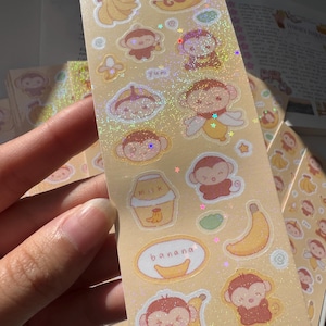 May include: A sheet of stickers with cartoon monkeys and bananas. The stickers have a glittery, holographic finish. Text includes "banana" and "milk". The color scheme is light yellow and brown. The stickers are designed with a cute, playful aesthetic.