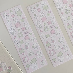 May include: Three sheets of pink and white stickers featuring cute cartoon bunnies holding four-leaf clovers. The stickers are decorated with small pink flowers and green clovers. The text on the stickers reads "&copy; 2024 dear mini!"