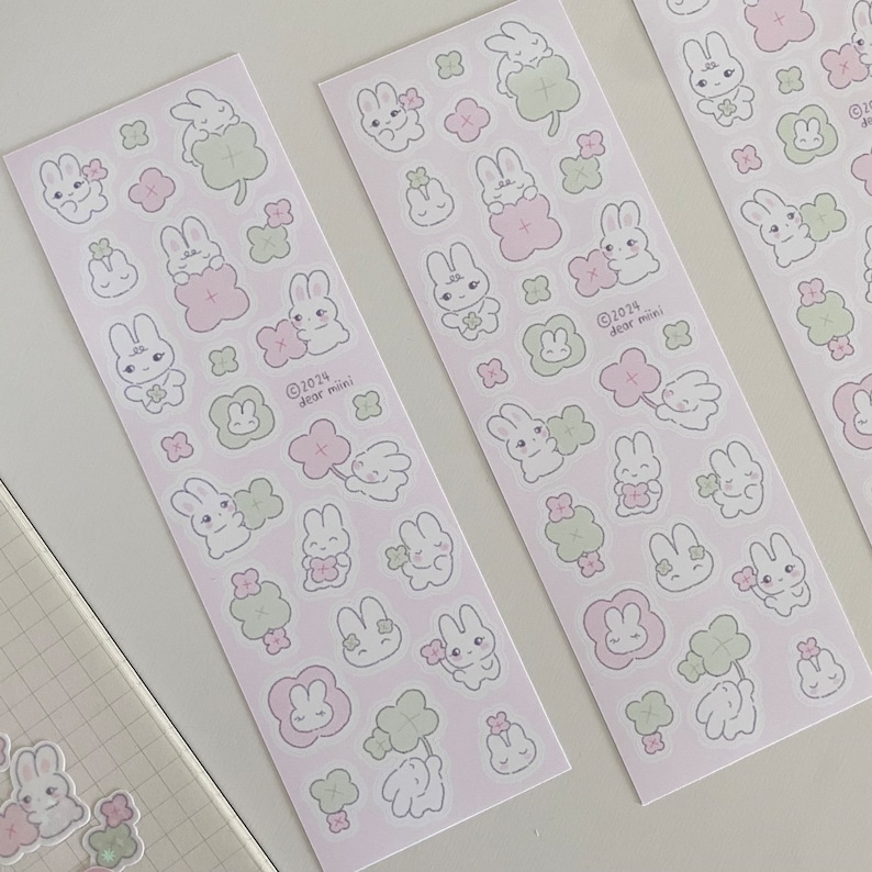 May include: Three sheets of pink and white stickers featuring cute cartoon bunnies holding four-leaf clovers. The stickers are decorated with small pink flowers and green clovers. The text on the stickers reads "&copy; 2024 dear mini!"