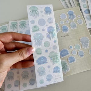 May include: A sheet of clear stickers with holographic jellyfish and other sea-themed designs. The stickers are on a white background and have a light blue and green color scheme. The text "Jellyfish" is written on the sheet.