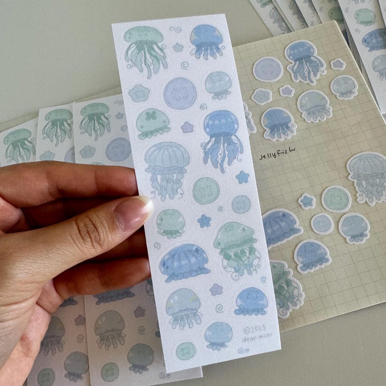 May include: A sheet of stickers featuring illustrations of jellyfish in various shades of blue and green. The stickers are on a white background with a few stars and other small details. The text "Jelly Fish" is written in the bottom right corner of the sheet.