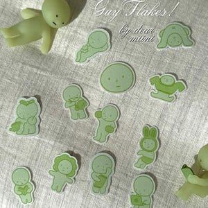 May include: A collection of light green stickers featuring various cartoon figures, including one lying down, one reading, and one with a laptop. The text reads Mini Green Guy Flakes! by dear miini. The stickers are approximately 4 cm in diameter.