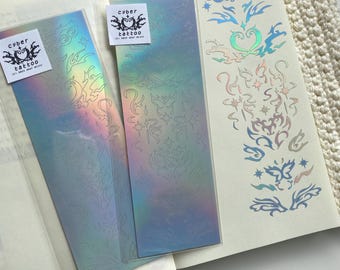 May include: Two holographic temporary tattoo sheets with a variety of designs, including butterflies, swirls, and floral patterns. The sheets are labeled "cyber tattoo" and are displayed on an open notebook.
