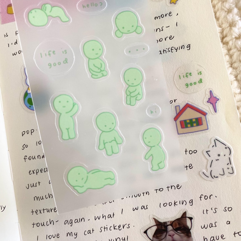 May include: A sheet of clear stickers with green cartoon figures in various poses. The stickers feature the text "life is good" and "hello?"