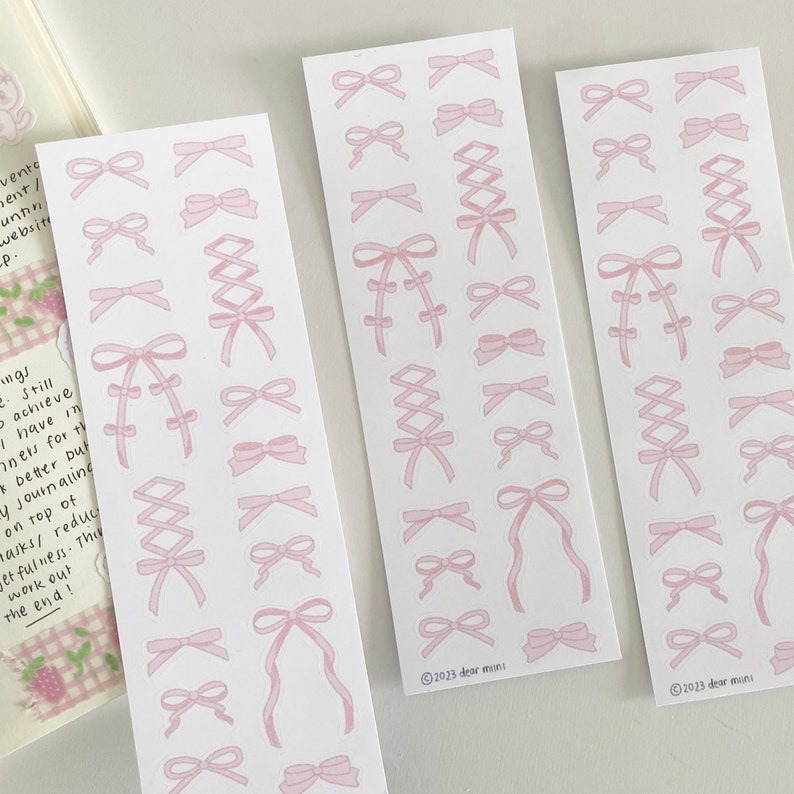 May include: Three sheets of pink and white sticker paper with various pink ribbon designs. The stickers feature bows, criss-cross ribbons, and other ribbon designs. The sheets are labeled with the text "2023 dear mini".