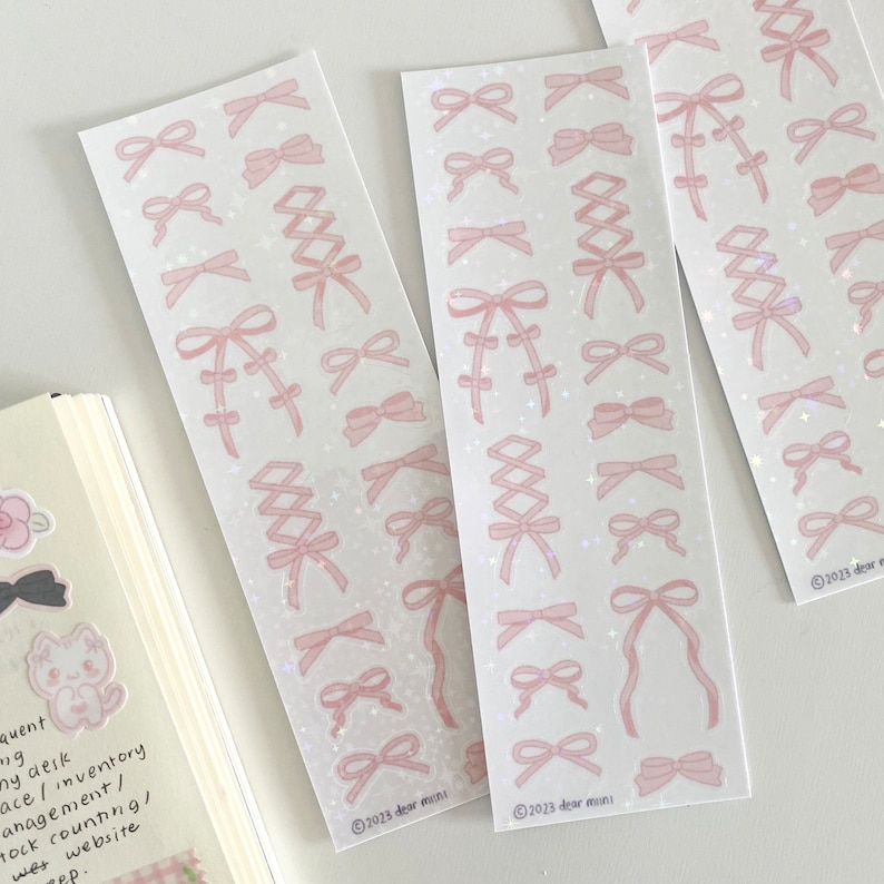 May include: Three sheets of pink and white sticker paper with various pink bow designs. The stickers are on a white background with a subtle glitter effect. The bottom of each sheet has the text "2023 dear mini".