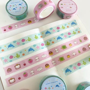 May include: Four rolls of washi tape with different designs. The designs include a pink and white pattern with bunnies, donuts, and cherries, a blue and white pattern with dolphins, seashells, and cacti, and a green and white pattern with dolphins, seashells, and cacti. The text on the tape reads "Ocean Treasure" and "Sweet Dream".