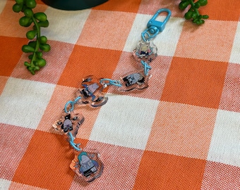 May include: A blue keychain with a series of clear plastic charms featuring cartoon characters. The charms are connected by a silver chain. The keychain is on a white and orange checkered surface.