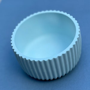 Può includere: Una ciotola in ceramica blu chiaro con un design scanalato e nervato. La ciotola è vuota e ha un interno liscio e arrotondato.
