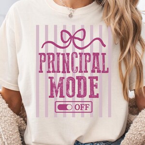 May include: Ivory-colored t-shirt with the text "PRINCIPAL MODE OFF" in a pink, distressed font. The design includes a bow and vertical stripes. The person is wearing denim shorts and a cream-colored cardigan.