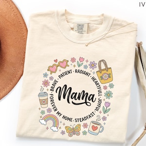May include: Ivory-colored t-shirt with a floral wreath design around the word "Mama" in black script. The wreath includes sunglasses, a coffee cup, a rainbow, a butterfly, and a handbag. The words "Brave, Patient, Radiant, Heartful, Joyful, Forever My Home, Steadfast" are also included.