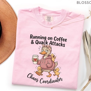 May include: Light pink t-shirt with the text "Running on Coffee & Quack Attacks" and "Chaos Coordinator". The graphic features a cartoon duck wearing a hoodie and holding a coffee cup, surrounded by ducklings.