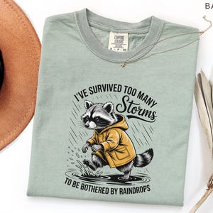 May include: A light green t-shirt featuring a raccoon wearing a yellow raincoat, walking through a puddle in the rain. The text reads, "I've survived too many storms to be bothered by raindrops."