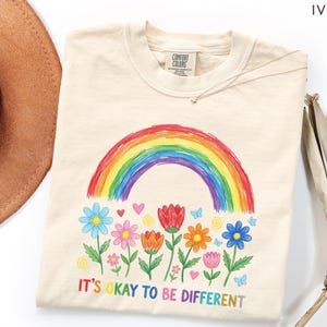 May include: Ivory-colored t-shirt with a colorful rainbow and floral design. The shirt features a rainbow arching over a row of flowers in various colors. The text "IT'S OKAY TO BE DIFFERENT" is printed below the flowers.