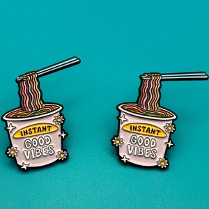 Könnte beinhalten: Zwei Emaille-Pins in Form von Instant-Nudelbechern mit herausstehenden Essstäbchen. Die Becher sind rosa mit einem gelben Etikett, auf dem "Instant Good Vibes" steht.