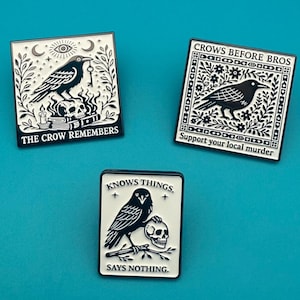 Könnte beinhalten: Drei quadratische Emaille-Anstecknadeln mit schwarz-weißen Raben-Illustrationen. Eine trägt die Aufschrift THE CROW REMEMBERS mit Totenkopf. Eine weitere zeigt CROWS BEFORE BROS und Support your local murder. Die dritte trägt den Text KNOWS THINGS. SAYS NOTHING.