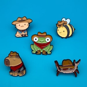 Könnte beinhalten: Fünf Emaille-Pins mit Cartoon-Tieren, die Cowboyhüte tragen. Die Pins zeigen eine Katze, einen Frosch, eine Biene, ein Capybara und eine Krabbe. Alle Pins befinden sich auf einem leuchtend blauen Hintergrund.