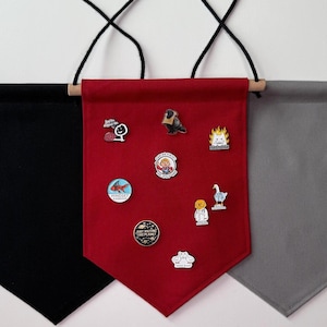 May include: Three fabric pennants hanging from a white wall. The pennants are black, red, and grey. The red pennant has several enamel pins attached to it.