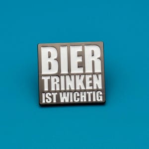 Könnte beinhalten: Eine rechteckige Emaille-Anstecknadel mit dunkelgrauem Hintergrund und fetter weißer Schrift, die BIER TRINKEN IST WICHTIG lautet. Das Design ist so strukturiert, dass das Wort BIER in der oberen Zeile, TRINKEN in der Mitte und IST WICHTIG unten steht.