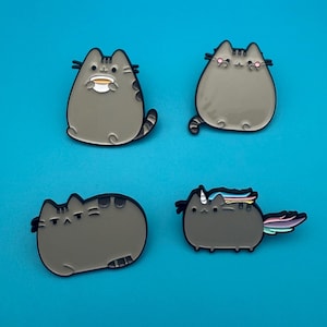 Könnte beinhalten: Vier graue Katzen-Emaille-Anstecknadeln mit schwarzen Umrissen. Eine zeigt eine Katze mit einer Tasse, eine andere eine Katze mit rosa Wangen, eine dritte eine grummelige Katze und die letzte eine Katze mit einem Regenbogen-Einhorn-Schwanz. Auf blauem Hintergrund.