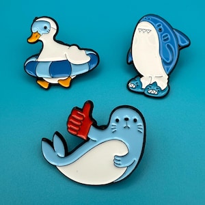 Könnte beinhalten: Drei Emaille-Pins mit einem Enten-Motiv, das eine Schwimmbrille und eine Schwimmhilfe trägt, einem Hai mit blauen Hausschuhen und einer Robbe mit einem roten Fäustling, die einen Daumen hoch zeigt.