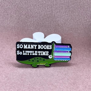 Könnte beinhalten: Ein grüner Alligator mit einem Stapel bunter Bücher im Maul. Der Alligator hält ein Schild mit der Aufschrift "So many books, so little time".
