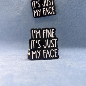 Könnte beinhalten: Zwei schwarze Emaille-Anstecknadeln mit gewelltem Rand, die jeweils den weißen Text "I'M FINE IT'S JUST MY FACE" in einer fetten, serifenlosen Schrift zeigen. Die Anstecknadeln sind vertikal angeordnet, eine direkt über der anderen platziert.