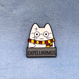 Könnte beinhalten: Weißer Emaille-Pin mit einer Katze, die einen rot-gelben Schal und eine Brille trägt. Die Katze hält einen Zauberstab und der Text "Expelliarmus" befindet sich unter der Katze.