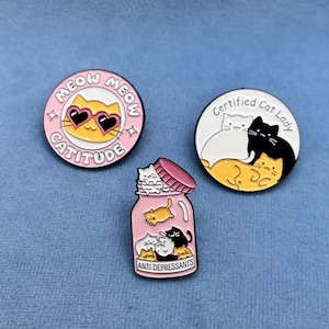 Könnte beinhalten: Drei Anstecknadeln aus Emaille mit Katzenmotiven. Eine Nadel ist rosa und trägt die Aufschrift "Meow Cattitude Meow". Eine weitere Nadel ist rosa und zeigt ein Glas mit Katzen darin und die Aufschrift "Anti Depressants". Die letzte Nadel ist weiß und trägt die Aufschrift "Certified Cat Lady" mit drei Katzen darin.