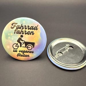 Könnte beinhalten: Ein runder Button mit dem deutschen Text "Fahrrad fahren ist veganes Reiten" und einer Fahrradillustration. Der Button hat einen bunten Hintergrund. Ein zweiter Button liegt daneben.