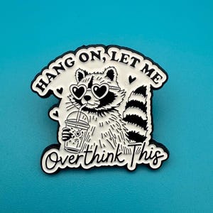 May include: A black and white enamel pin depicting a raccoon wearing heart-shaped sunglasses and drinking from a cup. The pin has the text "Hang on, let me overthink this."