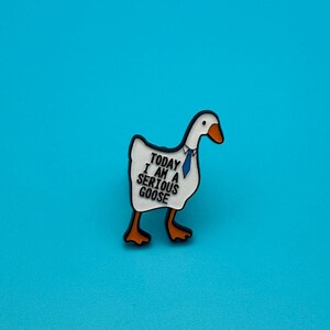 Könnte beinhalten: Ein weißer Gänse-Anstecker mit orangefarbenem Schnabel und Füßen, der eine blaue Krawatte trägt. Der Anstecker trägt den Text "TODAY I AM A SERIOUS GOOSE" in schwarzen Buchstaben. Der Hintergrund ist einfarbig blau.