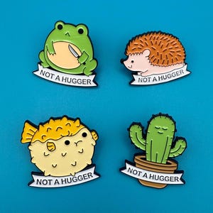 Könnte beinhalten: Vier Emaille-Pins mit Comic-Tieren und dem Text "Not a hugger". Die Pins zeigen einen grünen Frosch, der ein Messer hält, einen braunen Igel, einen gelben Kugelfisch und einen grünen Kaktus in einem braunen Topf.
