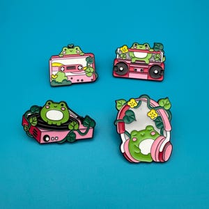Könnte beinhalten: Vier Emaille-Anstecknadeln mit Cartoon-Fröschen. Ein Frosch befindet sich auf einem rosa Kassettenband, ein anderer auf einem rosa Ghettoblaster, ein dritter auf einem rosa Plattenspieler und der letzte trägt einen rosa Kopfhörer. Jede Nadel hat grüne Blätter und gelbe Blumen.