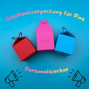 Könnte beinhalten: Drei farbige Geschenkboxen: rot, pink und blau, auf einem leuchtend blauen Hintergrund. Auf der pinkfarbenen Box steht "NAME". Der Text im Bild lautet "Geschenkverpackung für Pins" und "Personalisierbar".