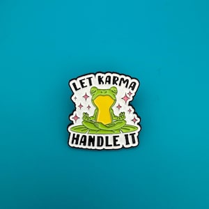 Könnte beinhalten: Ein skurriler Emaille-Pin mit einer meditierenden Frosch auf einem Seerosenblatt. Der Pin hat den Text "LET KARMA HANDLE IT" in einem Schwarz-Weiß-Design mit rosa Sternakzenten auf einem tealblauen Hintergrund.