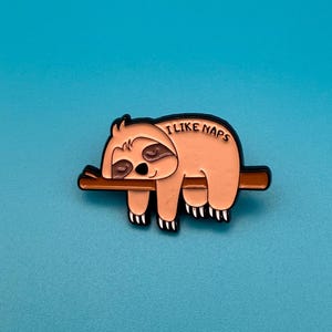 Può includere: Uno spillo in smalto nero e marrone con un bradipo dei cartoni animati che dorme su un ramo. Il bradipo è marrone chiaro e ha un naso e occhi neri. Il testo "I LIKE NAPS" è scritto sulla schiena del bradipo.