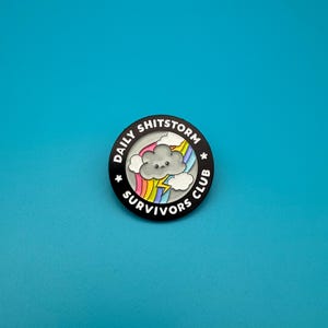 May include: Black enamel pin with a white outline. The pin features a cartoon cloud with a rainbow and lightning bolt. The text around the cloud reads "Daily Shitstorm Survivors Club".