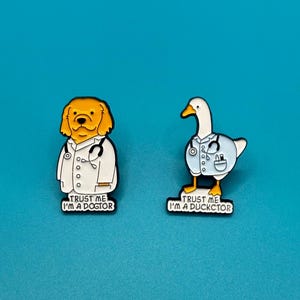 Peut inclure: Deux épingles en émail représentant un golden retriever et une oie habillés en médecins. Les épingles disent "Trust me I'm a Dogtor" et "Trust me I'm a Ducktor".