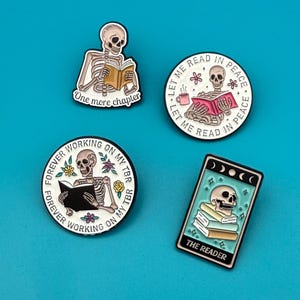 May include: Four enamel pins featuring skeletons reading books. The pins are white with black outlines and feature colorful details. The pins say "One more chapter", "Let me read in peace", "Forever working on my TBR", and "The Reader".