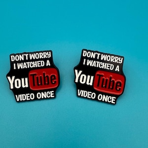 Può includere: Due spille in smalto nero con testo bianco che recita "Non preoccuparti, ho visto un video di YouTube una volta". La parola "YouTube" è in rosso e bianco.