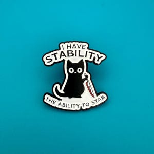 Può includere: Spilla a forma di gatto nero con la scritta "I HAVE STABILITY" e "THE ABILITY TO STAB". Il gatto tiene in mano un coltello insanguinato. La spilla è bianca con dettagli neri e rossi.