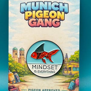 Op de afbeelding: Een kleurrijke pin met een rode vis met een haaienvin, geplaatst op een lichtblauwe cirkel. De pin bevat de tekst "MINDSET IS EVERYTHING" en "MUNICH PIGEON GANG". De achtergrond toont een stadsgezicht.
