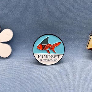 Könnte beinhalten: Eine blaue und weiße Emaille-Anstecknadel mit einem roten und weißen Fisch, der eine Haiflosse trägt. Der Text auf der Anstecknadel lautet "Mindset is Everything".