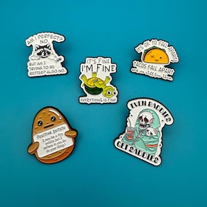 Op de afbeelding: Een verzameling emaille pins met humoristische illustraties en teksten. Ontwerpen omvatten een wasbeer, schildpad, taco, aardappel en skelet. Teksten zijn onder meer "Am I perfect? No" en "Even baddies get saddies."