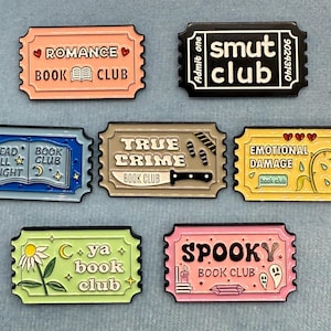 Könnte beinhalten: Satz von zehn Emaille-Pins mit Buchclub-Motiven. Die Pins sind in Form von Ticket-Stubs gestaltet und zeigen verschiedene Designs, darunter ein Buch mit einem Halbmond, ein Messer, eine Rose und ein Geist. Die Pins sind alle in verschiedenen Farben, darunter Blau, Gelb, Pink, Grün und Braun.